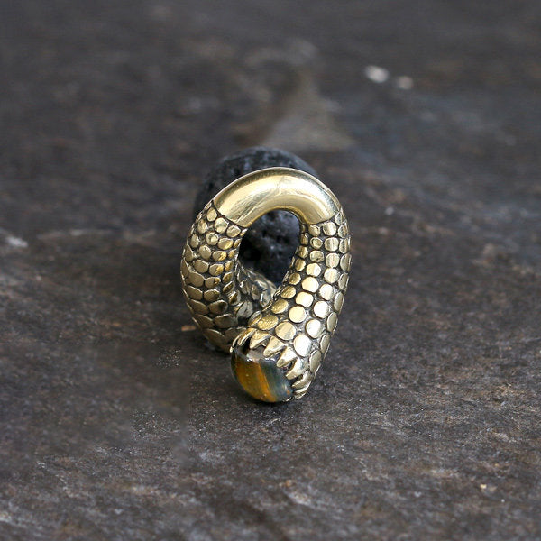A pair of TIMO Ear Weights in Gold with Tiger Eye by Kaya Movement rests on a dark, textured surface. These ear weights feature intricate, scaled detailing and are partially oxidized, giving them an aged, antique appearance. The head of each weight has a small, polished tiger's eye stone inset.